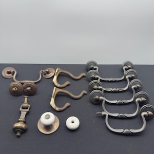 Może przedstawiać: Kolekcja antycznych okuć do szafek, w tym uchwyty do szuflad, gałki i haczyki. Okucia mają różne wzory i wykończenia, w tym mosiądz i metal. Uchwyty do szuflad mają zakrzywiony kształt, a gałki są z białej ceramiki.