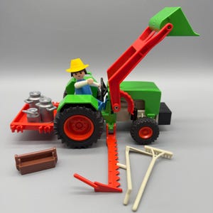 Puede incluir: Un tractor de juguete verde y rojo con cargador frontal, conducido por una pequeña figura con sombrero amarillo. Incluye un remolque rojo con latas de leche plateadas, una caja de herramientas marrón y herramientas agrícolas.