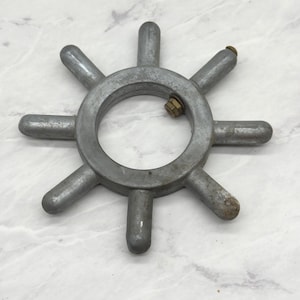 Puede incluir: Una rueda de metal de color plateado con ocho radios espaciados uniformemente que se extienden desde un cubo circular central. Una tuerca y un perno de color latón aseguran una parte de la rueda. La rueda es probablemente un componente de un dispositivo mecánico más grande.