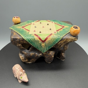 May include: A ceramic miniature game board with a green and red design, set on a textured brown base. Two small orange spheres sit on the sides. A small, detailed armadillo figurine is in the foreground.