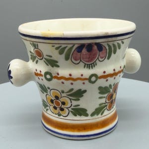 May include: A white ceramic shaving mug with a flared shape and two side handles. The mug is decorated with colorful floral patterns in orange, yellow, blue, and green. A blue and brown band encircles the base.