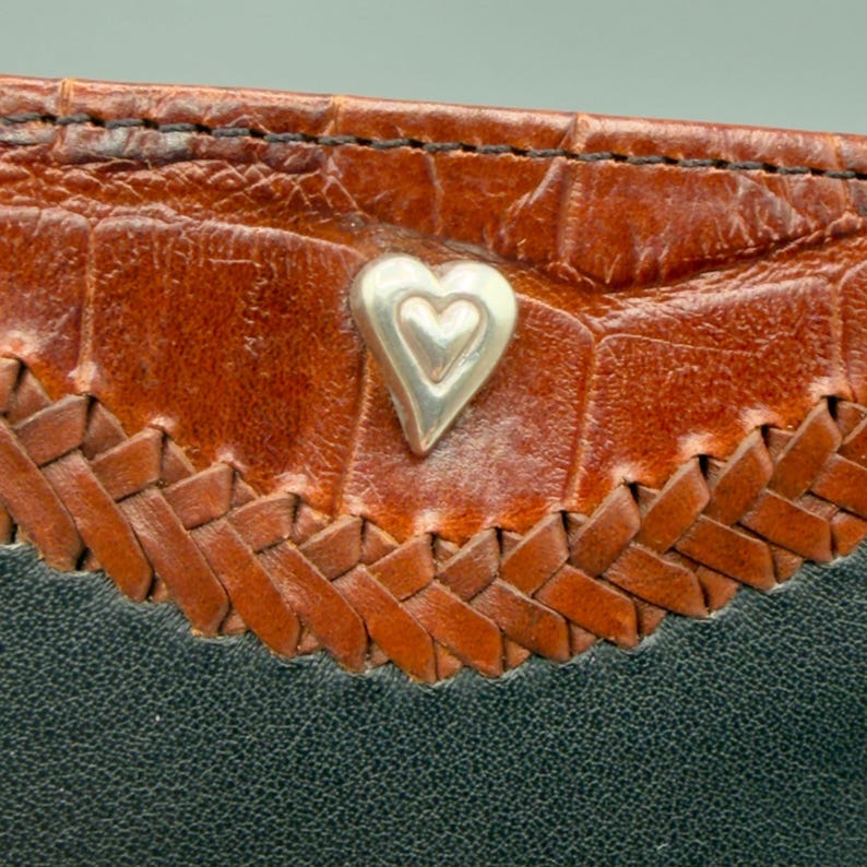 May include: Close-up of a leather item with a silver heart-shaped embellishment. The item features a textured brown leather section, a woven brown leather border, and a black leather base. The stitching is visible.