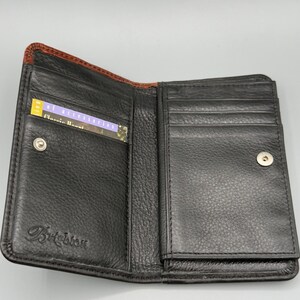 May include: Open black leather wallet with a brown trim. The wallet has multiple card slots and a snap closure. A card with the text "Classic Accent" is visible inside the wallet. The brand name "Brighton" is embossed on the front.