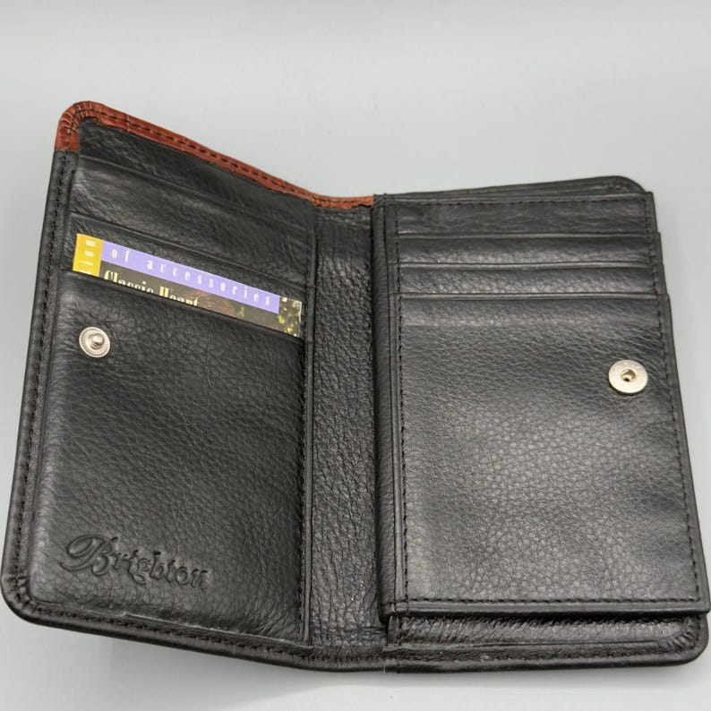 May include: Open black leather wallet with a brown trim. The wallet has multiple card slots and a snap closure. A card with the text "Classic Accent" is visible inside the wallet. The brand name "Brighton" is embossed on the front.