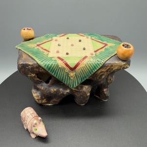 May include: A ceramic miniature game board with a green and red design, set on a textured brown base. Two small orange spheres sit on the sides. A small, detailed armadillo figurine is in the foreground.