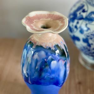 Puede incluir: Un jarrón de cerámica con un borde acampanado y ondulado que presenta un esmalte rosa suave. El cuerpo del jarrón muestra un vibrante efecto de esmalte goteado, que transita de un azul cobalto profundo en el centro a un azul sólido más claro en la base.