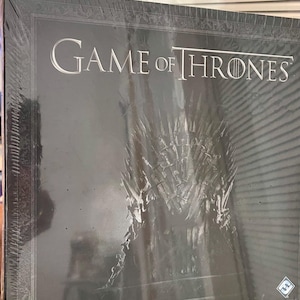 Może przedstawiać: Czarne pudełko z grą planszową Gra o Tron. Tytuł jest srebrnymi literami, z wizerunkiem Żelaznego Tronu. Logo HBO znajduje się w lewym dolnym rogu, z tekstem "A Game of Intrigue, Betrayal, and Struggle for Power."