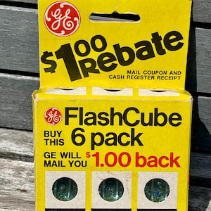 May include: A vintage yellow GE FlashCube 6-pack box. The box features the GE logo, the text "$1.00 Rebate", and the words "FlashCube 6 pack". The box contains six cubes, each with a green flashbulb. The text "6 Cubes/24 Guaranteed Flashes" is also visible.