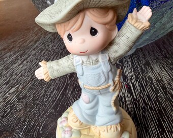 Może przedstawiać: Porcelanowa figurka stracha na wróble w zielonym kapeluszu z szerokim rondem, koszuli w kratę i jasnoniebieskich ogrodniczkach. Postać stoi na okrągłej podstawie ozdobionej małymi różowymi kwiatkami i żółtymi kamieniami, z rozłożonymi ramionami.