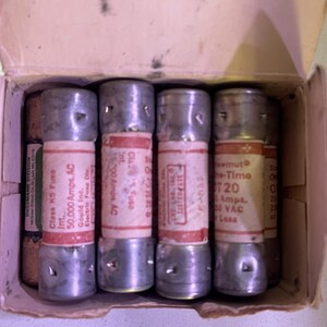 May include: A collection of cylindrical fuses with metal end caps and red and white labels. The labels contain text such as "Class K5 Fuse" and "50,000 Amps. AC". The fuses are stored in a cardboard box.