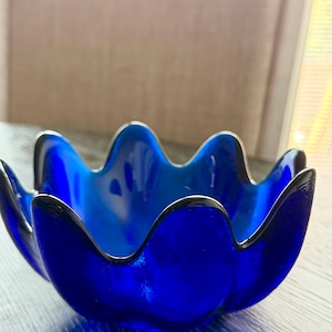 Pode incluir: Uma taça de vidro azul cobalto profundo com uma borda ondulada e festonada distinta. A taça tem um acabamento liso e translúcido com uma base arredondada, sendo que a cor azul vibrante parece mais saturada ao longo das bordas curvas.