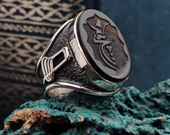 May include: A silver ring with a black onyx stone featuring Arabic calligraphy. The ring is set on a textured rock.