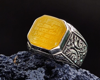 May include: A silver ring with an ornate design and a yellow stone set in the center. The stone has Arabic script carved into it.