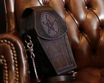 May include: A black leather coffin-shaped bag with a purple pentagram design and a silver chain strap. The bag is sitting on a brown leather chair.