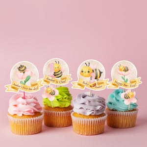 Puede incluir: Cuatro cupcakes con glaseado colorido y adornos decorativos. Cada adorno presenta una ilustración de acuarela de una abeja y una flor, con el texto "Amber's First Bee-day!", "Amber is One!", "Baby Amber" y "Happy Bee-Day!" sobre un fondo rosa.