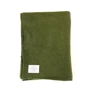 May include: Olive green wool blanket, folded neatly. A white tag with black text is visible on the blanket. The blanket is made of a thick, textured material. The blanket is a solid color.