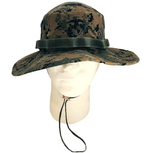 May include: A camouflage boonie hat with a wide brim, featuring a green and brown digital pattern. The hat has a green fabric band and a chin strap. An emblem is visible on the front of the hat.