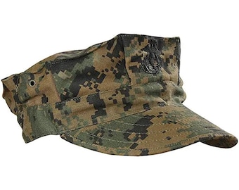 Puede incluir: Una gorra militar de camuflaje verde y marrón con un emblema bordado en negro de un ancla y un águila. La gorra tiene una visera rígida y un botón en el lateral.