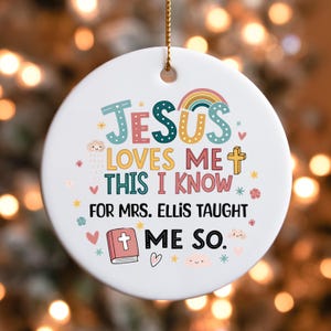 May include: A white, round Christmas ornament with the text "JESUS LOVES ME THIS I KNOW FOR MRS. ELLIS TAUGHT ME SO." in colorful letters. The ornament features a rainbow, cross, hearts, stars, and a book illustration.