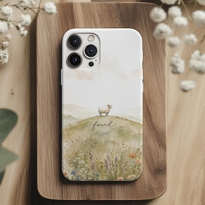 May include: A white smartphone case featuring a watercolor illustration of a small sheep standing on a grassy hill covered in wildflowers. The word found is written in a cursive script below the sheep. The case has a triple camera cutout.
