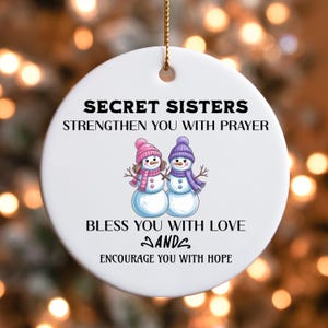 May include: A round white ceramic ornament featuring a gold hanging string. The text reads: SECRET SISTERS STRENGTHEN YOU WITH PRAYER BLESS YOU WITH LOVE AND ENCOURAGE YOU WITH HOPE. Between the lines is an illustration of two snowmen wearing hats.