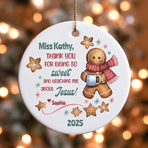 May include: A white ceramic Christmas ornament with a gold string. The ornament features a gingerbread man wearing a red scarf and holding a blue mug. Text reads "Miss Kathy, THANK YOU FOR BEING SO sweet and teaching me about Jesus!" and "Sophia 2025".
