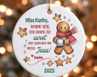May include: A white ceramic Christmas ornament with a gold string. The ornament features a gingerbread man wearing a red scarf and holding a blue mug. Text reads "Miss Kathy, THANK YOU FOR BEING SO sweet and teaching me about Jesus!" and "Sophia 2025".