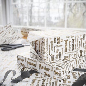 May include: Gift boxes wrapped in white paper with gold religious text, including "Savior" and "Immanuel." A roll of matching paper, scissors, and a gray ribbon are also visible, suggesting a holiday or special occasion.