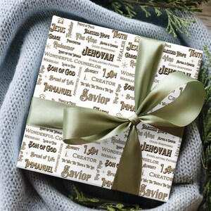 May include: A wrapped gift with white paper featuring religious words in a repeating pattern, including "Jesus" and "Savior." The gift is tied with a sage green satin ribbon and bow, creating a festive presentation.