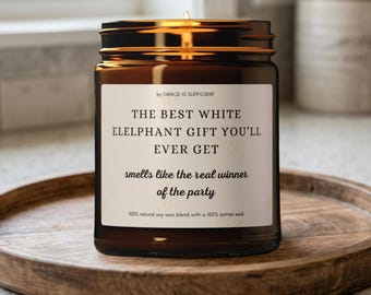 Könnte beinhalten: Eine braune Glas-Kerze mit Metalldeckel, auf einem Holztablett. Das Etikett lautet "THE BEST WHITE ELELPHANT GIFT YOU'LL EVER GET" und "smells like the real winner of the party."