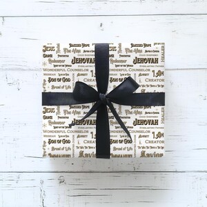 May include: A wrapped gift with a black ribbon and bow. The wrapping paper is white with gold text featuring religious words such as "Jesus", "Jehovah", and "Savior". The gift is on a white wooden surface.