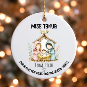 May include: A white ceramic Christmas ornament with a gold string. The ornament features a Nativity scene with the text "Miss Tanya" and "Thank you for teaching me about Jesus!" The ornament is personalized with "From, Lilah 2025".