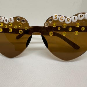 May include: Brown heart-shaped sunglasses with decorative clear and yellow rhinestones. The text "THE SCRIPT" is spelled out in white letter beads across the top of the lenses. The sunglasses have brown frames and lenses.