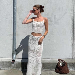 May include: A white crochet skirt and top set with a brown crocheted bag. The woman is wearing sunglasses and a hair clip.
