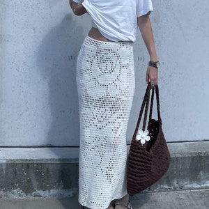 May include: A woman wearing a white t-shirt with the text "Something for everyone" printed on it, a white crocheted skirt, brown sandals, and a brown crocheted tote bag with a white flower.