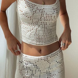 May include: A white crochet top and skirt set. The top is a sleeveless crop top with a scoop neck. The skirt is a midi skirt with a high waist.