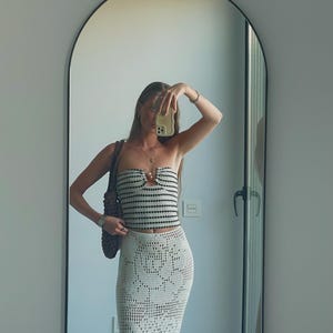 May include: A woman wearing a white and black striped crochet top with a crocheted white skirt. She is holding a phone in front of her face and has a brown bag slung over her shoulder.