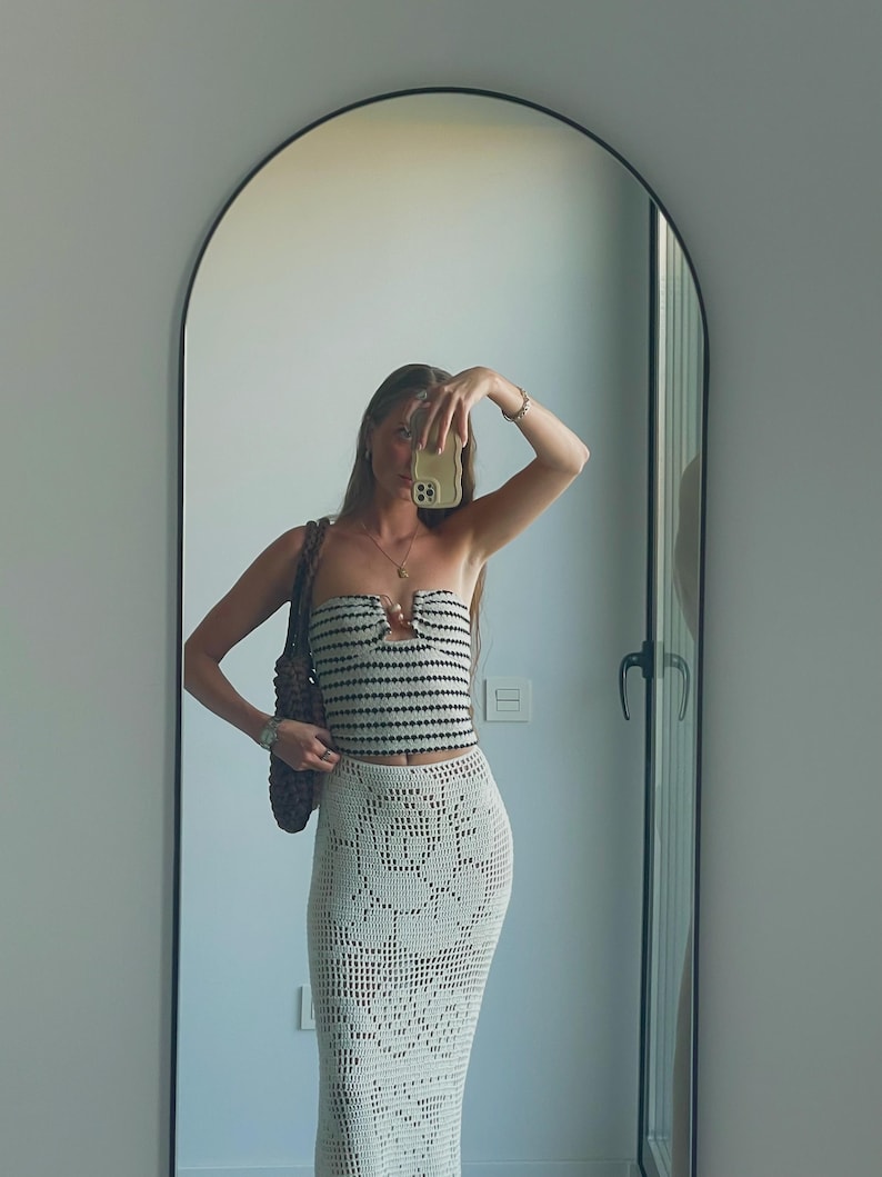 May include: A woman wearing a white and black striped crochet top with a crocheted white skirt. She is holding a phone in front of her face and has a brown bag slung over her shoulder.
