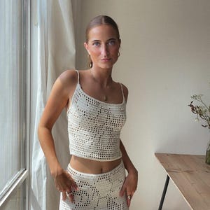May include: A white crochet top and skirt set. The top is a cropped style with a scoop neck and thin straps. The skirt is a midi length with a high waist.