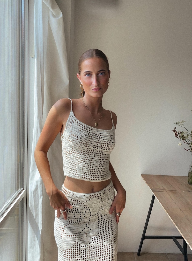 May include: A white crochet top and skirt set. The top is a cropped style with a scoop neck and thin straps. The skirt is a midi length with a high waist.
