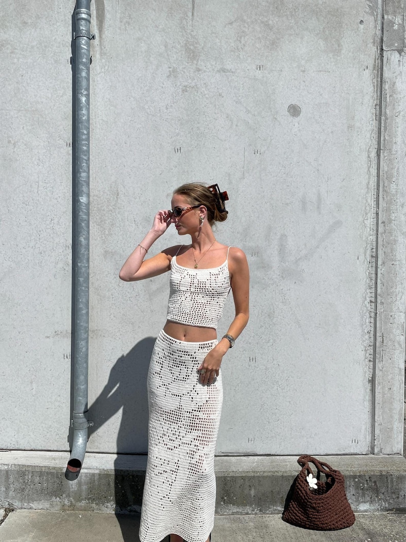 May include: A white crochet skirt and top set with a brown crocheted bag. The woman is wearing sunglasses and a hair clip.