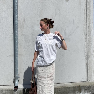May include: A woman wearing a white t-shirt with the text "SUPER COOL" printed on it, a white crochet skirt, and a brown crocheted tote bag.