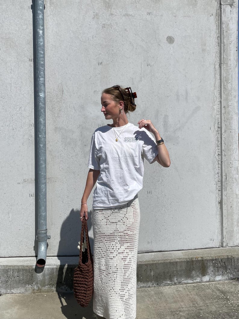 May include: A woman wearing a white t-shirt with the text "SUPER COOL" printed on it, a white crochet skirt, and a brown crocheted tote bag.