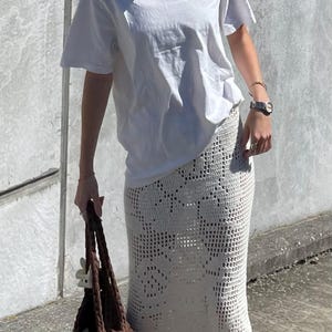 May include: A white t-shirt with the text "SPORTS & GLORY" printed on the front, paired with a white crochet skirt. The person is holding a brown crocheted tote bag.