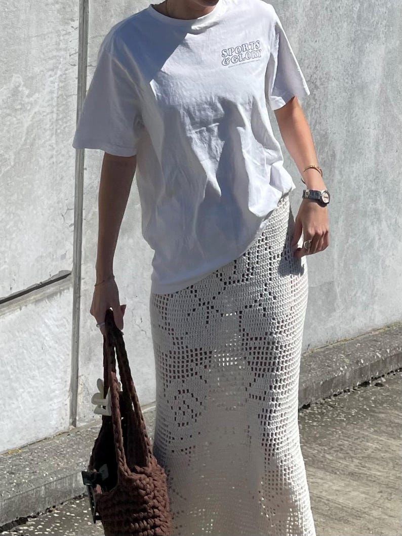 May include: A white t-shirt with the text "SPORTS & GLORY" printed on the front, paired with a white crochet skirt. The person is holding a brown crocheted tote bag.