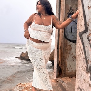 May include: A white crochet top and matching skirt with a delicate floral pattern. The top has thin straps and a scoop neck. The skirt is long and flowing.