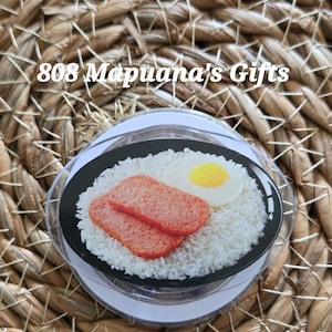 May include: A circular magnet featuring a graphic of a plate of white rice topped with two slices of pink luncheon meat and a fried egg with a yellow yolk. The text 808 Mapuana's Gifts is printed in white across the top of the circular design.