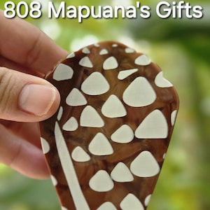 Puede incluir: Un objeto decorativo marrón y blanco con un patrón único de formas blancas sobre un fondo marrón. El objeto está sostenido por una mano y tiene el texto "808 Mapuana's Gifts" en la parte superior.