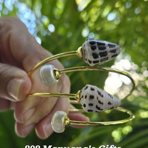 May include: Gold bangle bracelets with white and brown seashells. The bracelets are stacked on top of each other. 808 Mapuana's Gifts.