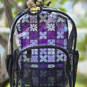 May include: Clear backpack with a purple floral pattern on the interior. The backpack has a black trim, mesh side pockets, and a front zippered pocket. The backpack is hanging from a tree branch.